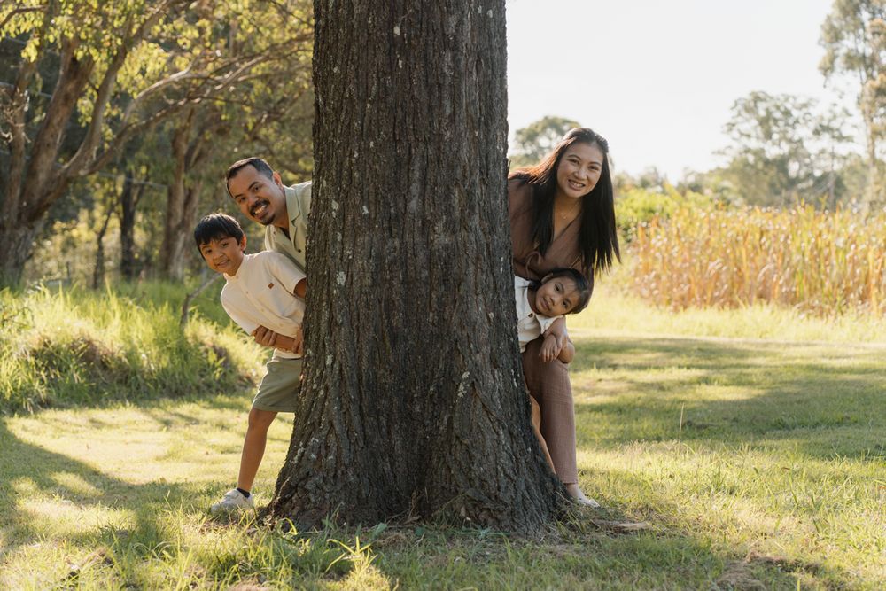 Family of four playfully peeking around a large tree trunk in a sunny green park setting during a family photoshoot in Rouse hill Sydney