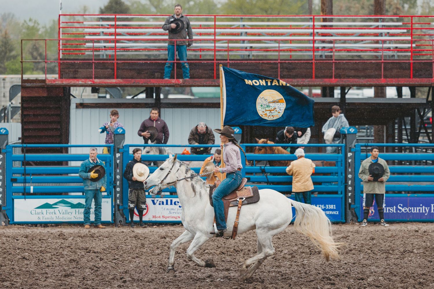 Cowboy rides white horse carrying Montana state flag at outdoor rodeo arena with red bleachers.