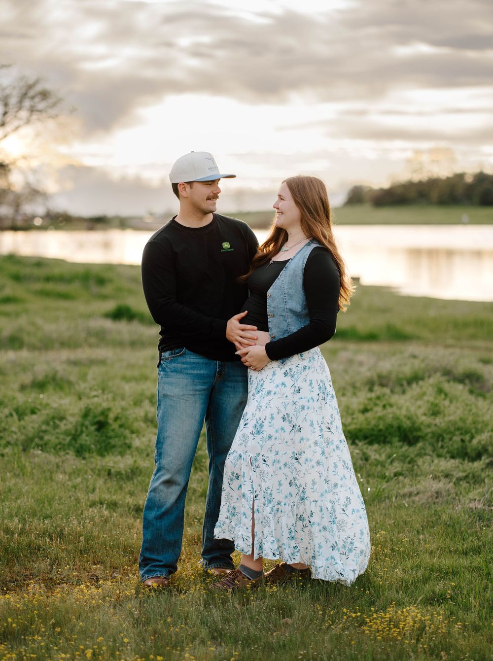 Couple smiling at each other in a green field at sunset, man in black sweater and cap, woman in floral dress.