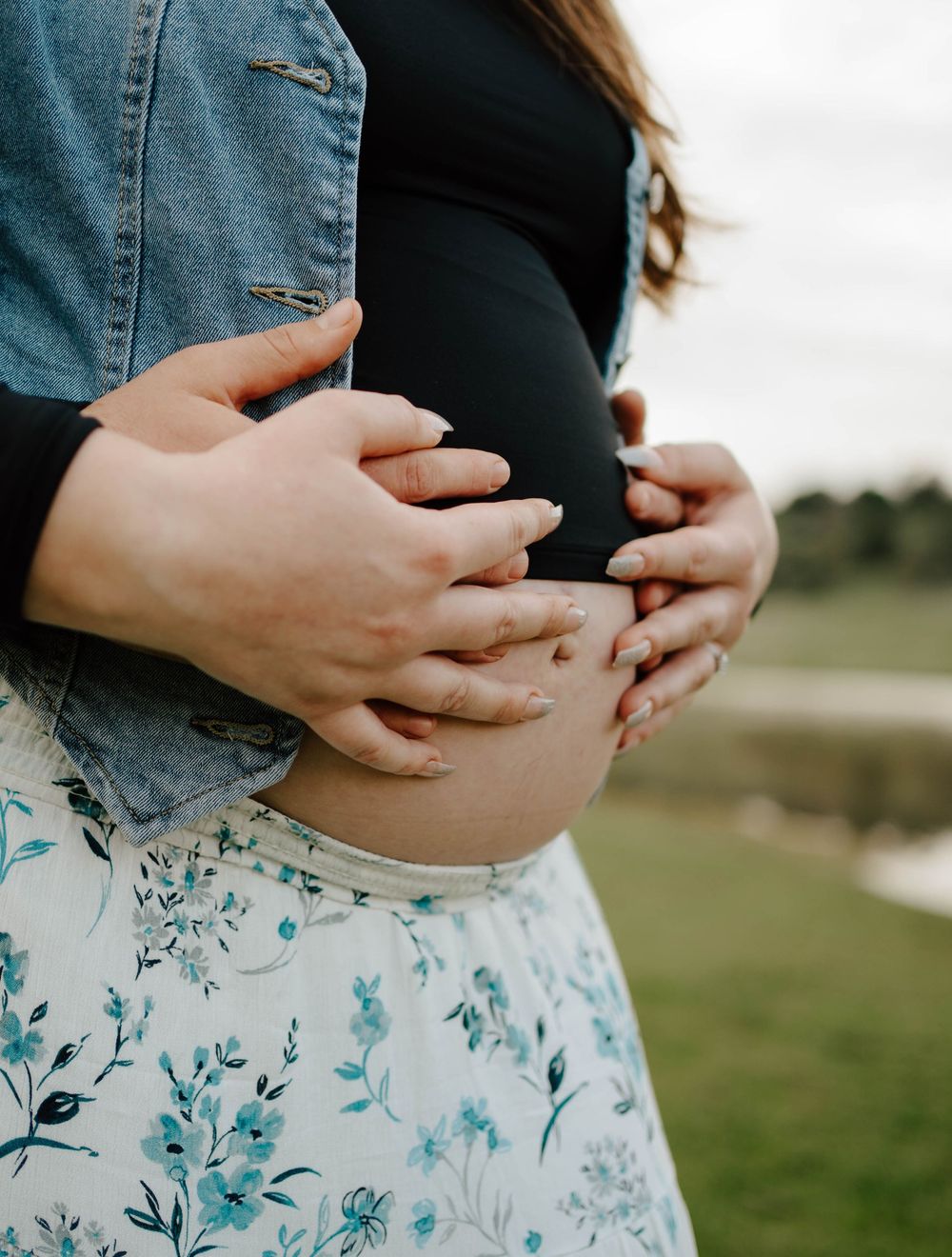 Couple cradling pregnant belly outdoors, woman in floral skirt and black top, partner in denim jacket.