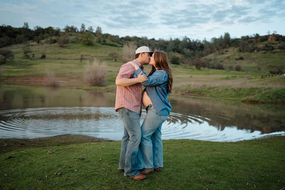 Couple sharing a romantic kiss by a serene pond with green hills in the background during an outdoor engagement session.