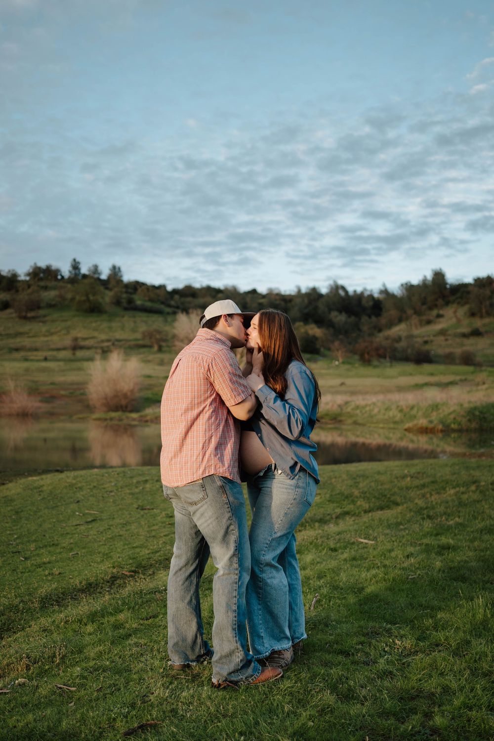 Couple sharing a kiss outdoors on green grass near a pond, man in pink shirt and woman in denim, golden hour light.