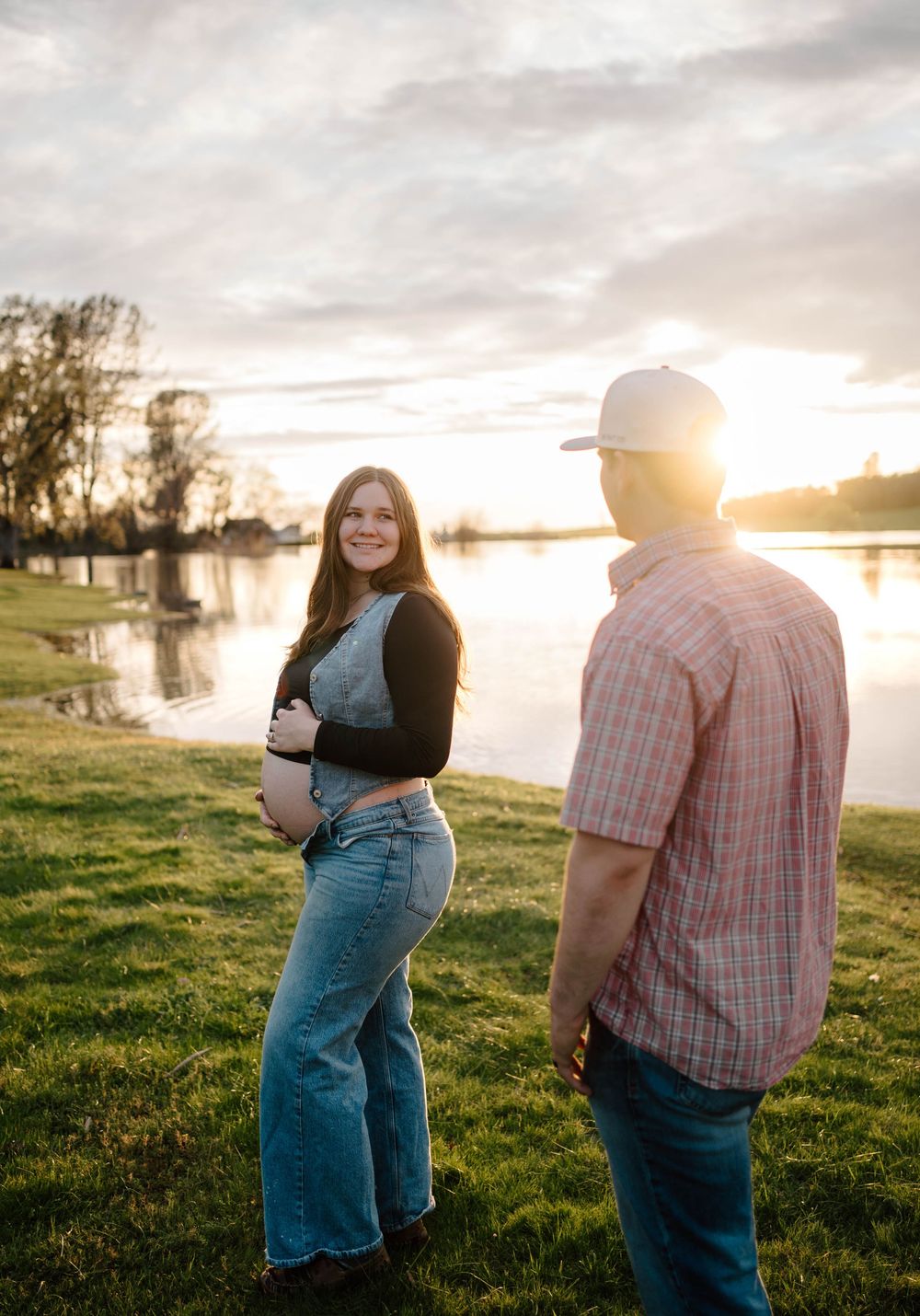 Pregnant woman smiling at sunset by a lake, standing with a man in a plaid shirt on green grass.