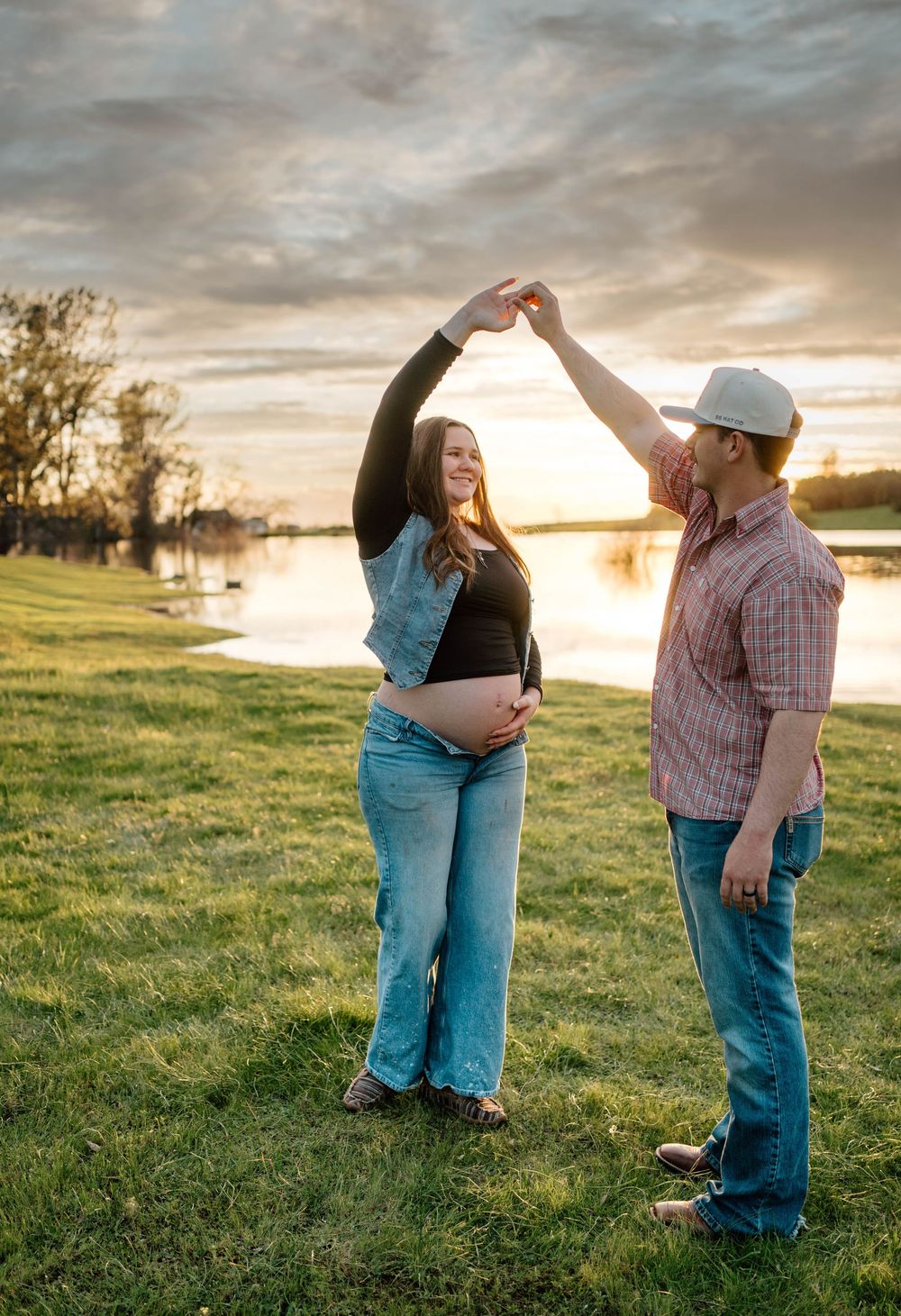 Pregnant couple dancing at sunset by a lake, man in plaid shirt twirling woman in jeans and black top on green grass.