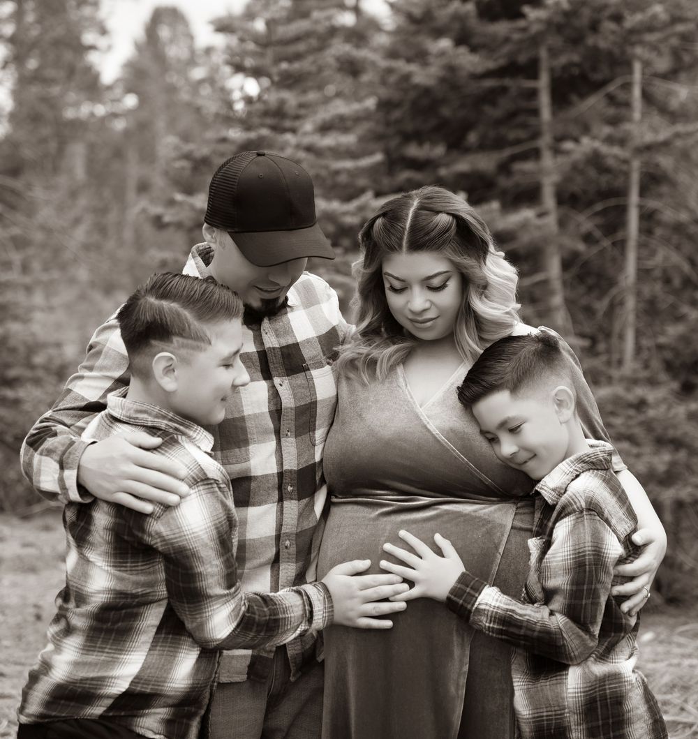 Black and white maternity photo of a family with two boys hugging pregnant mother outdoors near pine trees.