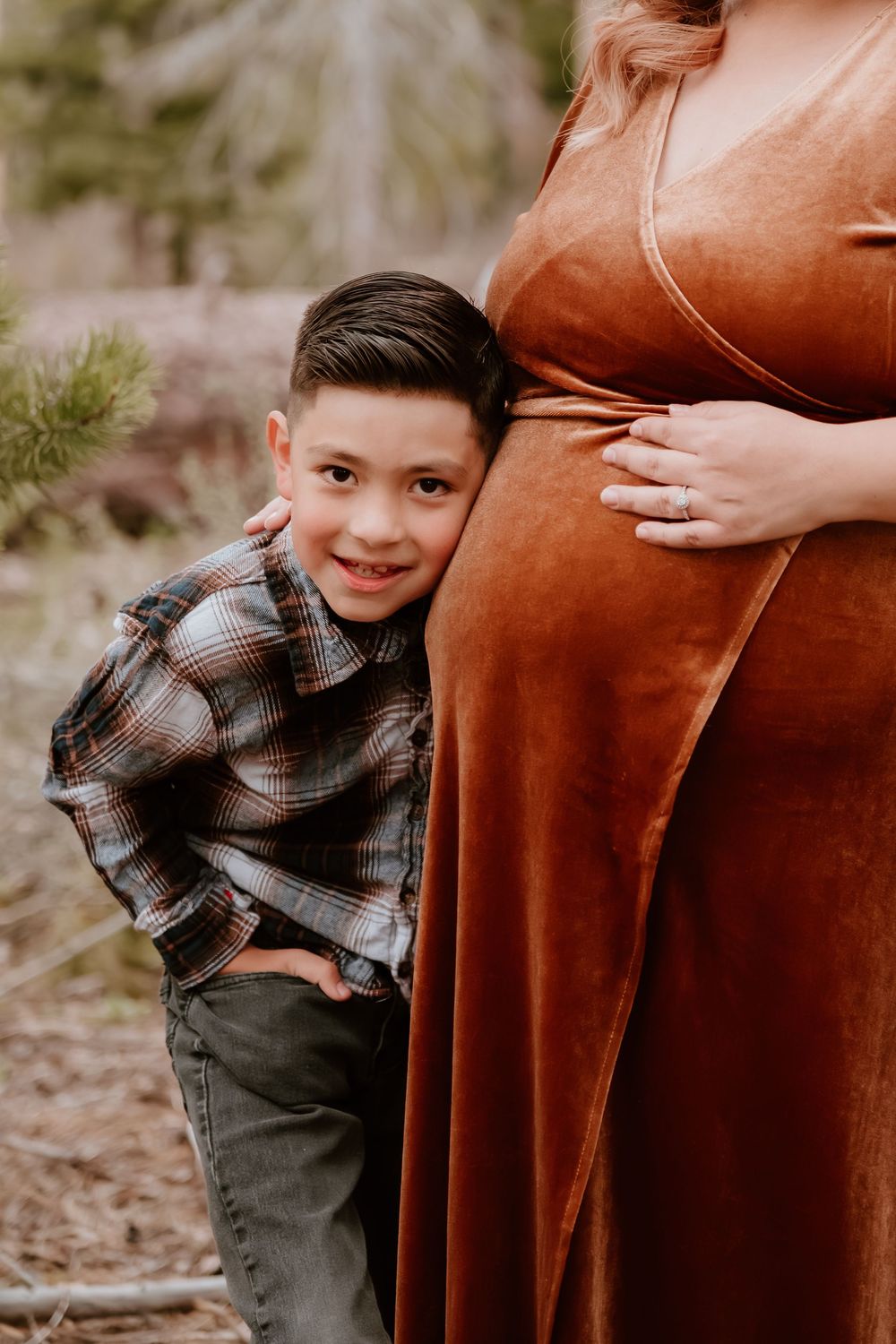 A young boy in a plaid shirt smiles while leaning against a pregnant woman in a rust velvet dress outdoors.