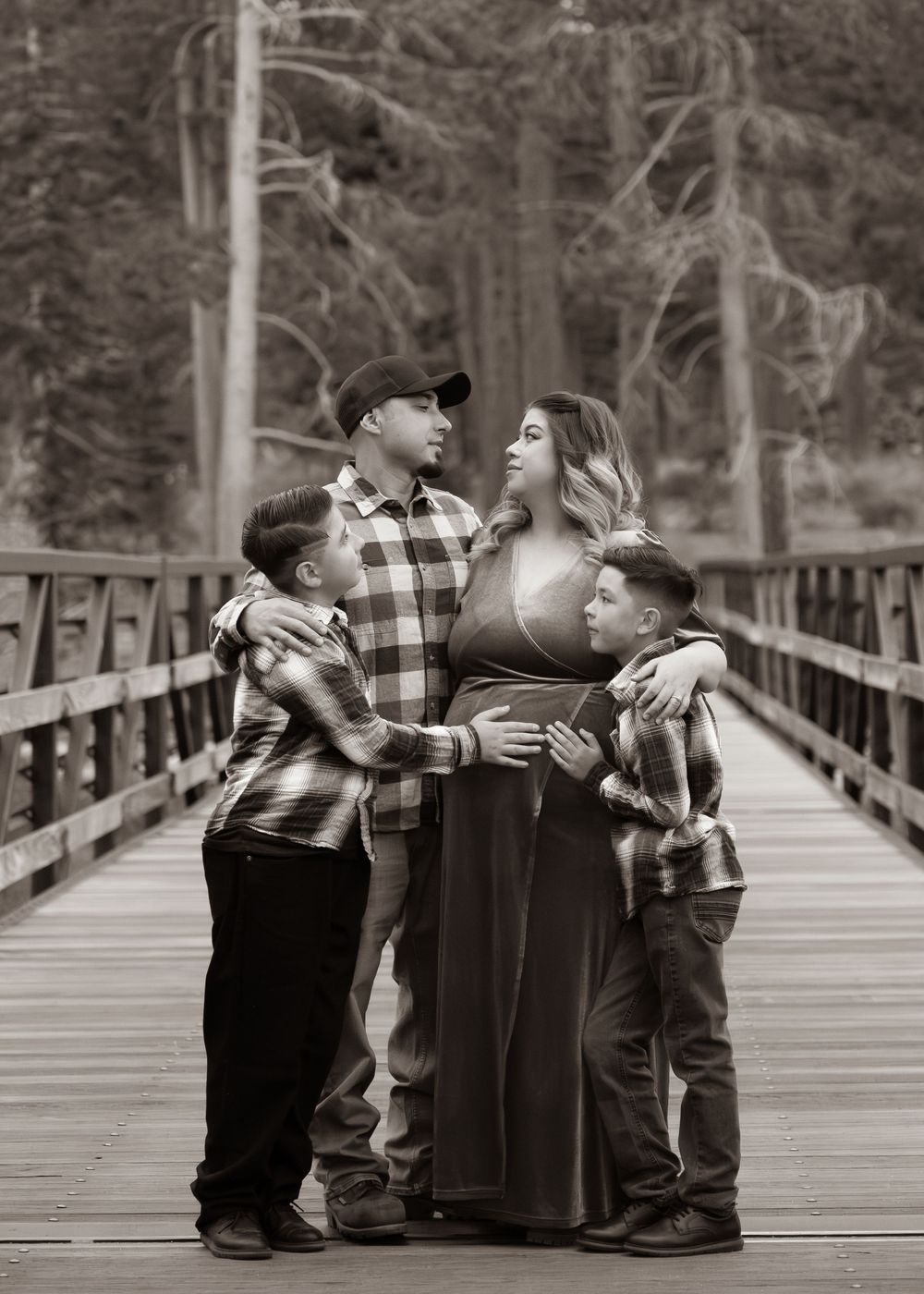 Black and white maternity photo of a family of four on a wooden bridge surrounded by tall trees.
