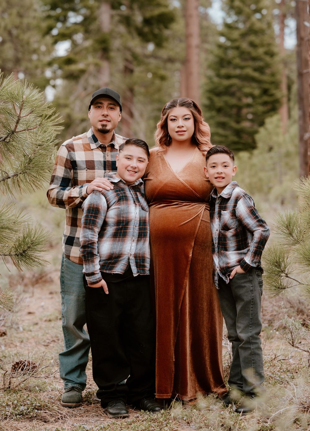 A pregnant woman in a rust velvet gown poses with her family among pine trees for a maternity photo shoot.
