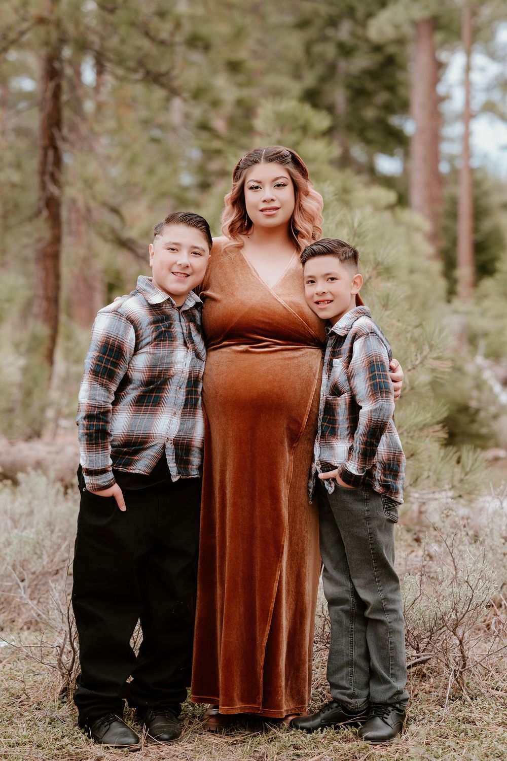 Pregnant woman in rust velvet dress poses with two boys in plaid shirts during outdoor maternity photo session.