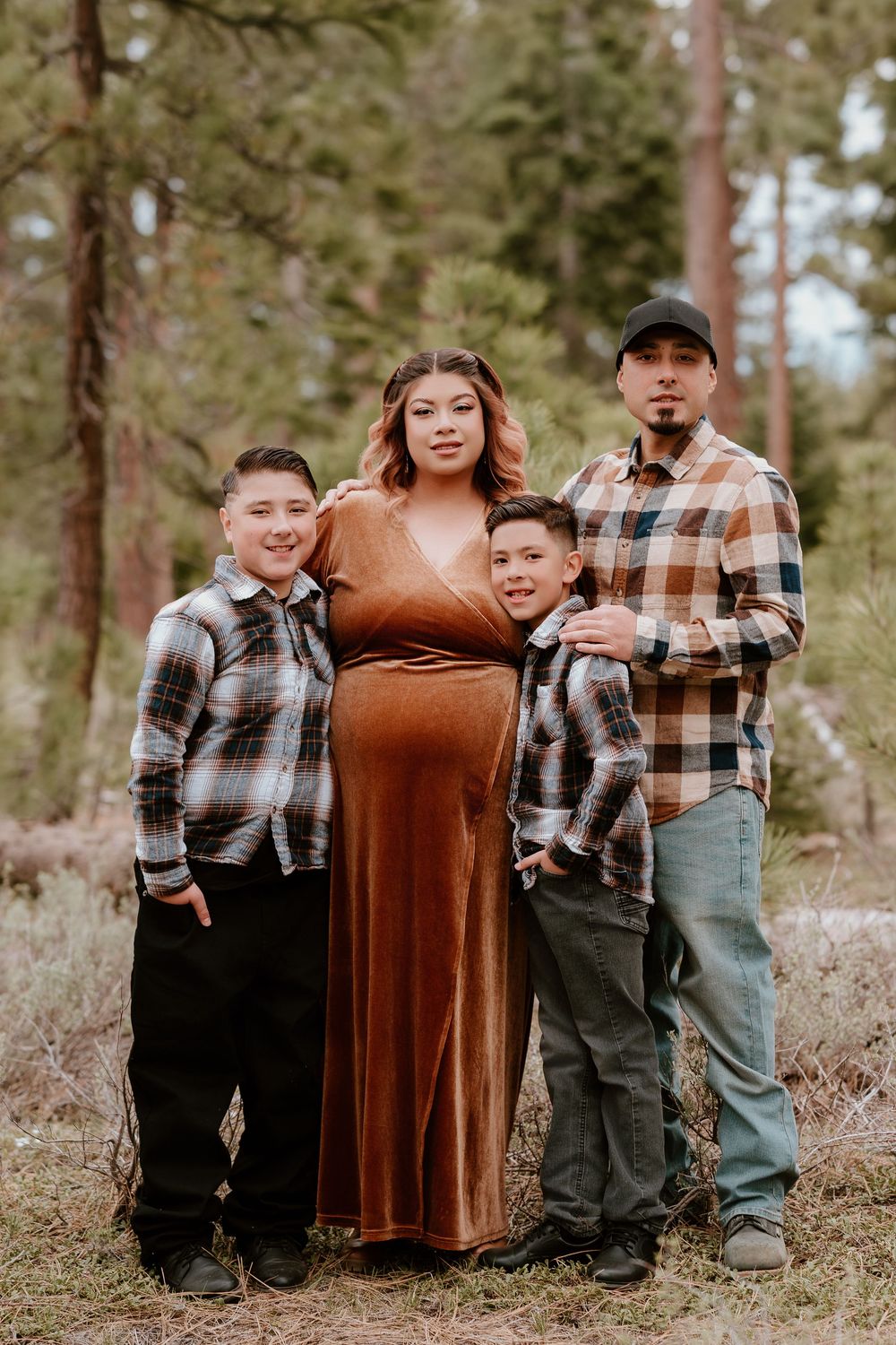Pregnant woman in rust dress poses with husband and two sons in plaid shirts for maternity photos in pine forest.