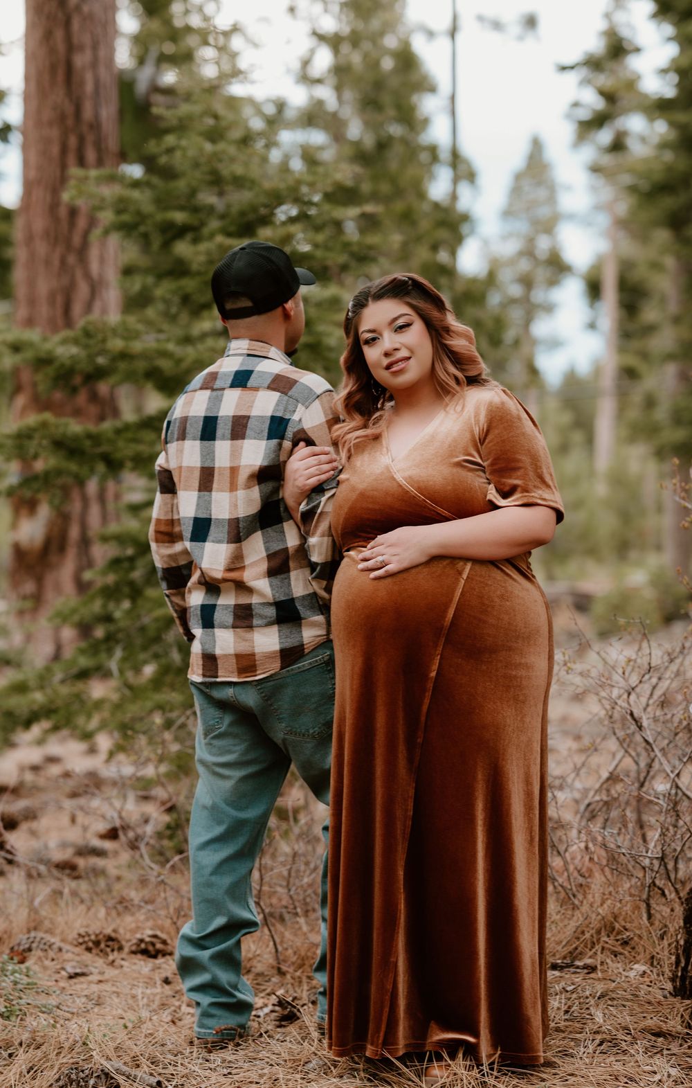 Pregnant woman in rust velvet gown poses with partner in plaid shirt during maternity photoshoot in pine forest.