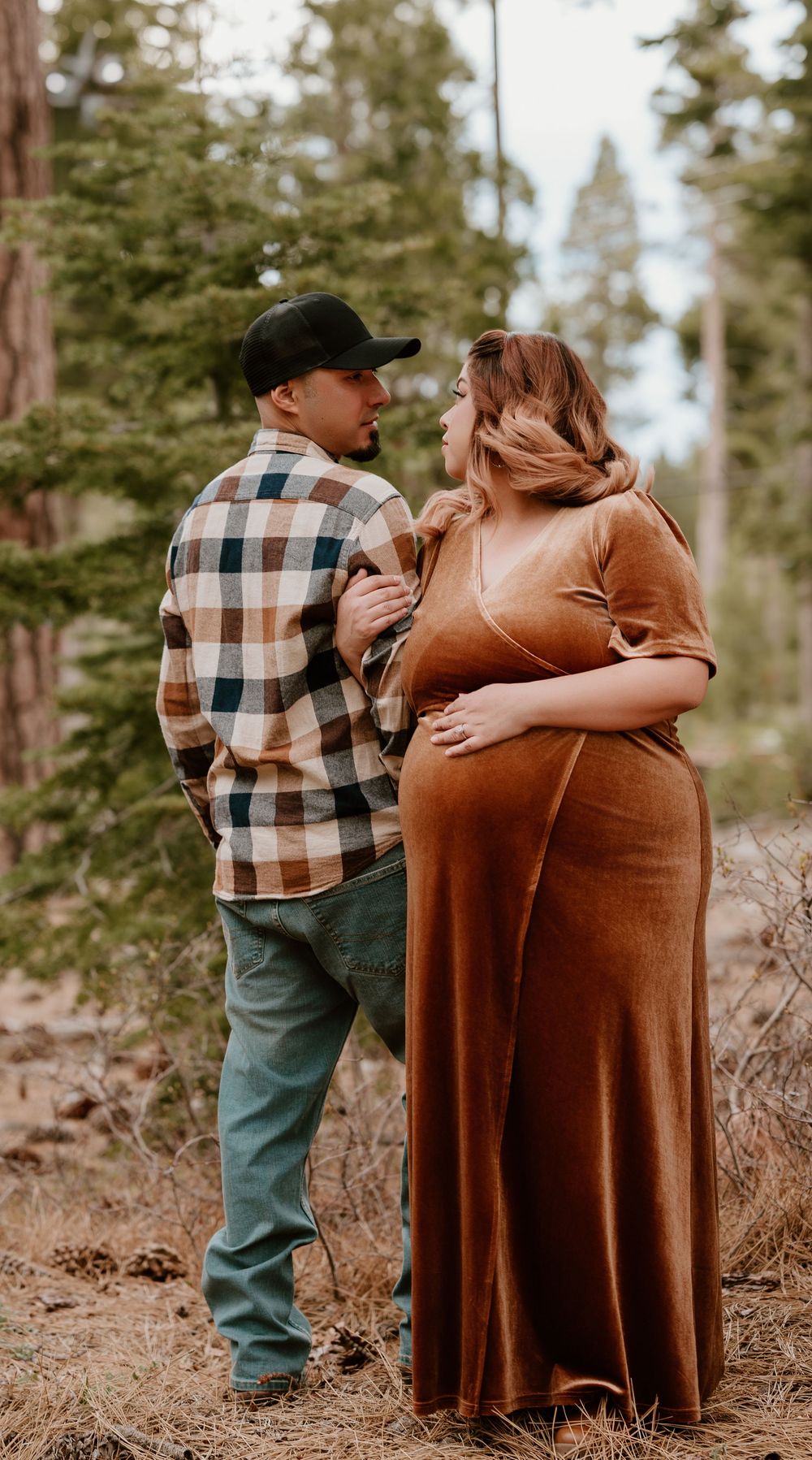 Couple shares tender moment outdoors, pregnant woman in rust velvet dress with partner in plaid shirt among tall pines.