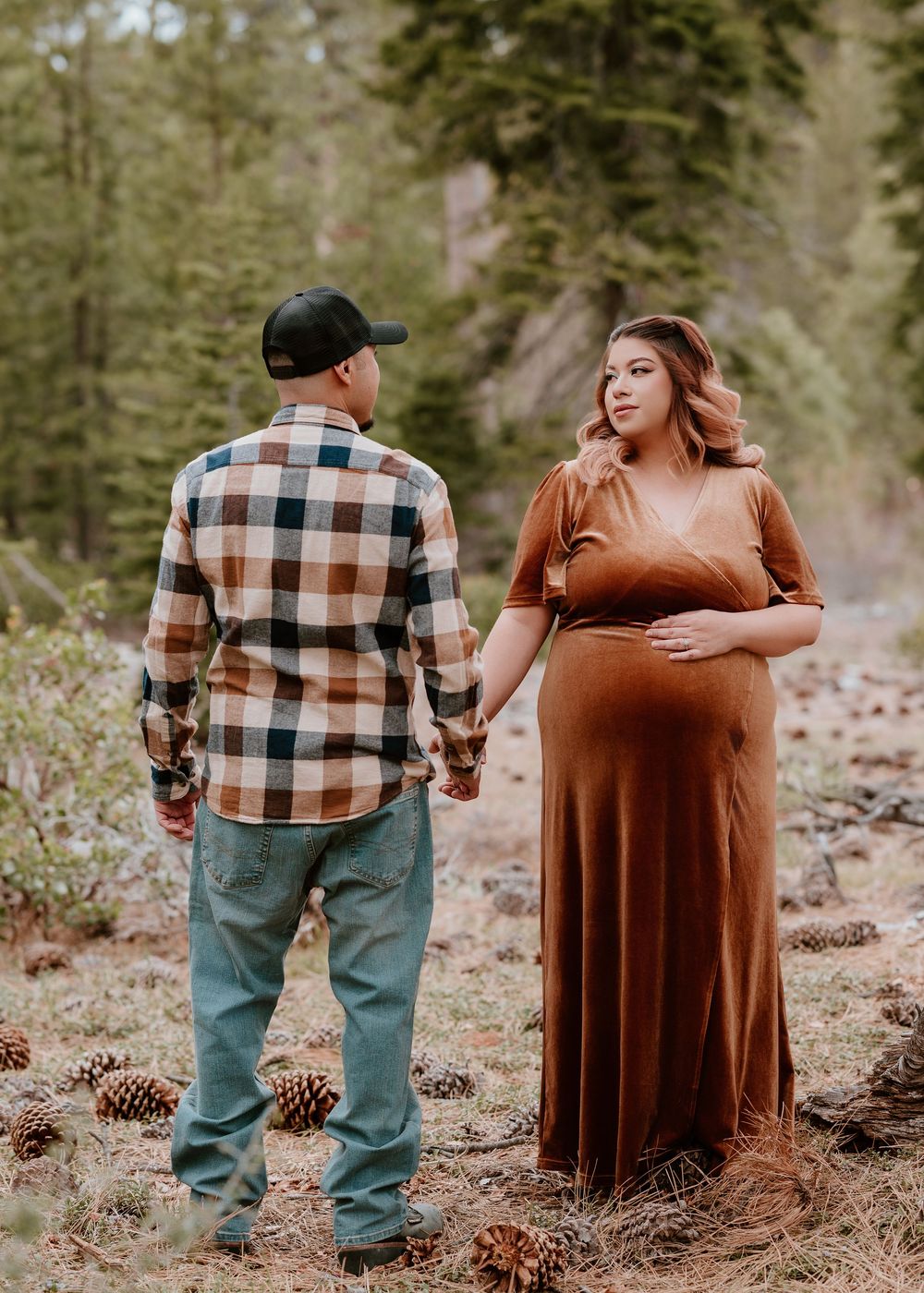 Pregnant woman in rust dress holds hands with man in plaid shirt amid pine trees for maternity photos.