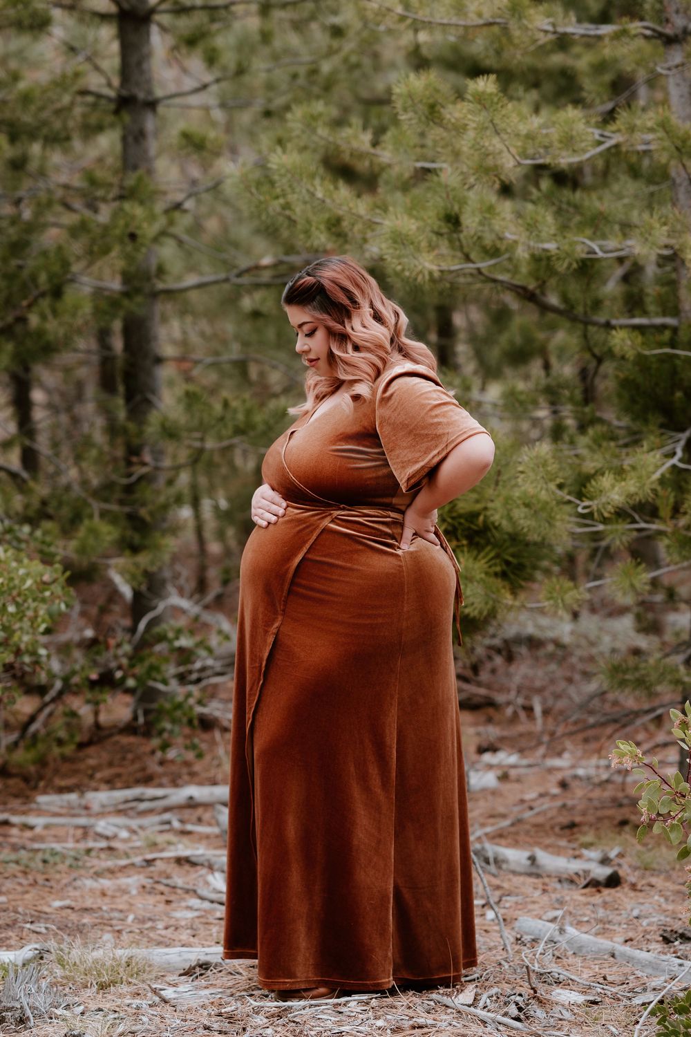 Pregnant woman in rust brown maxi dress standing in pine forest, cradling baby bump during outdoor maternity photo session.