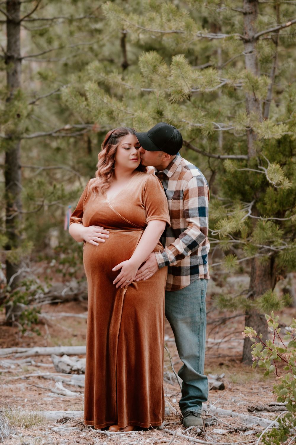 Pregnant woman in rust velvet gown kissed by partner in plaid shirt during outdoor maternity photoshoot in pine forest.
