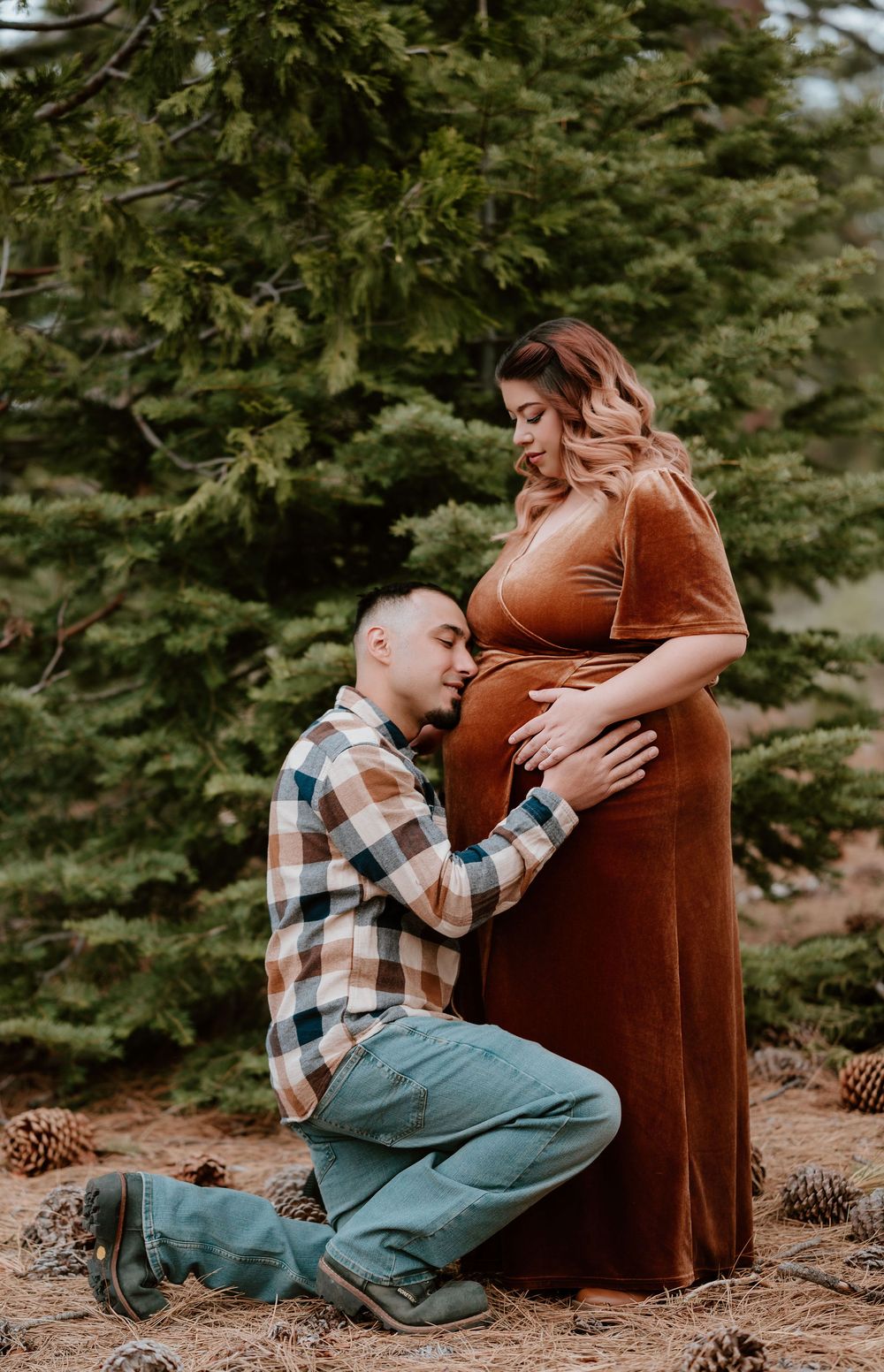 A man kneels kissing a pregnant woman's belly outdoors near pine trees, both dressed in warm autumn tones.