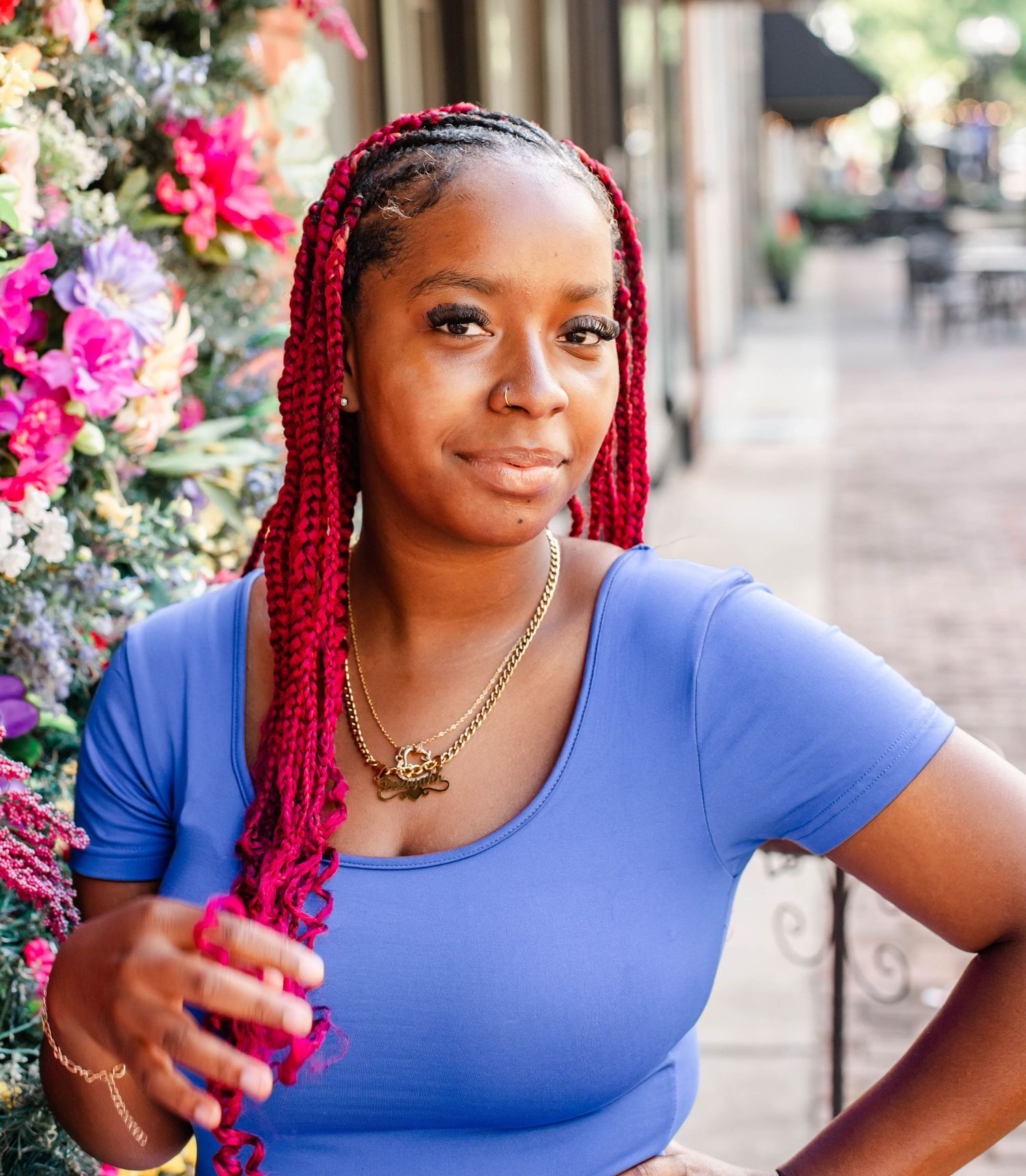 Young Black woman with red braids wearing a blue top and gold necklace posing near colorful flowers outdoors.
