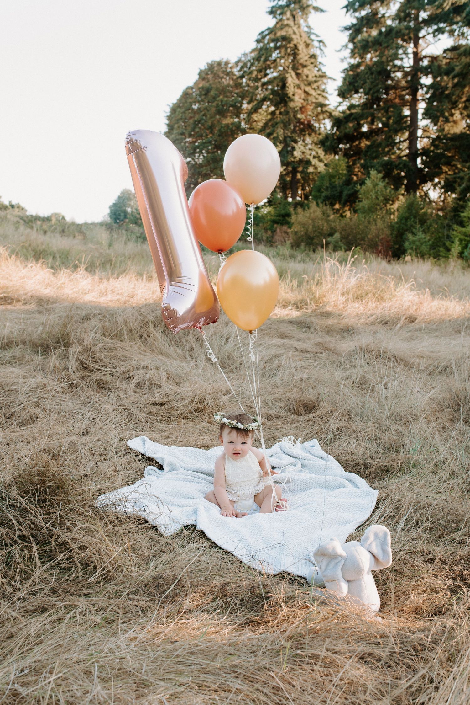 Baby girl sits on white blanket outdoors holding colorful balloons and a number 1 balloon for her first birthday.