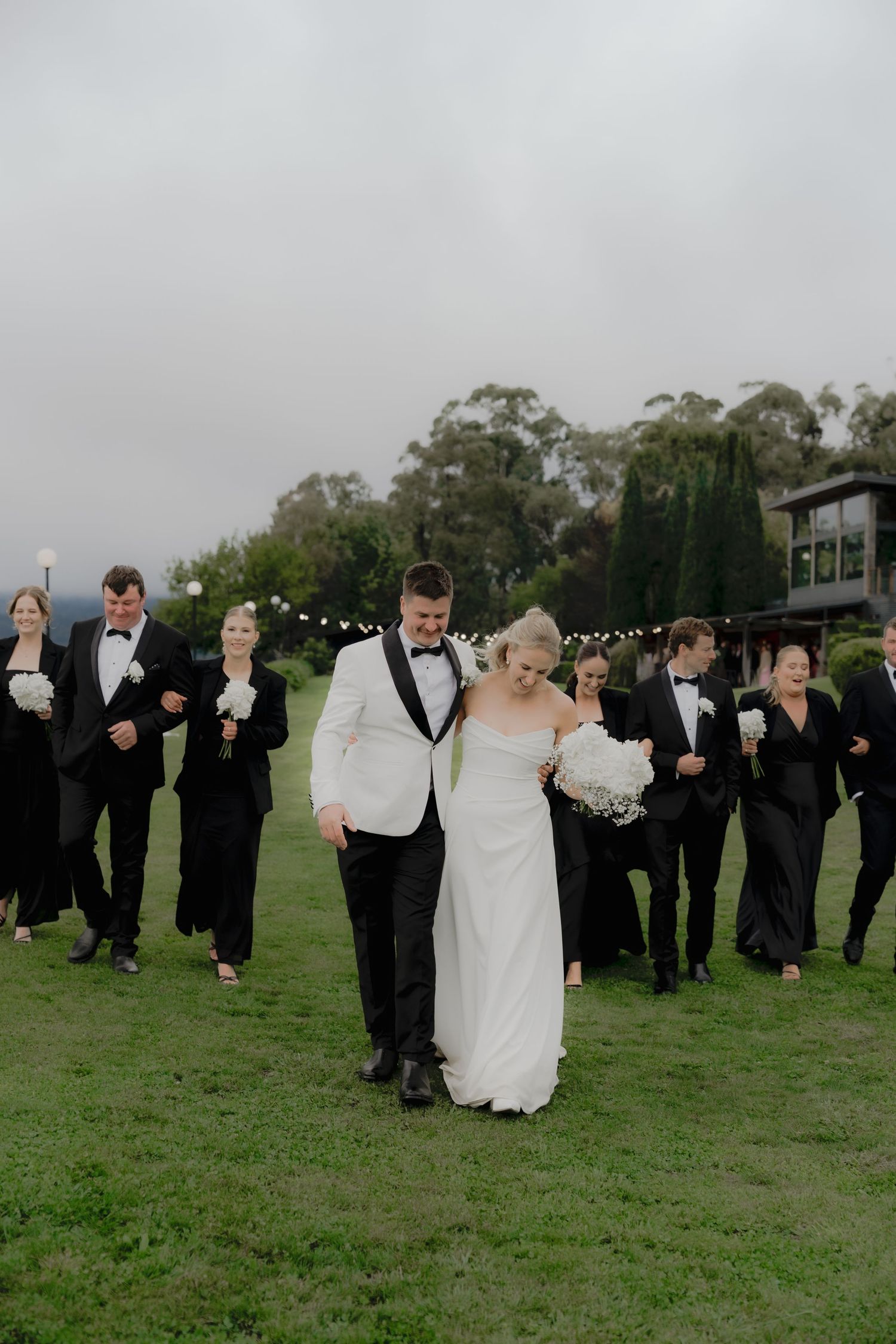 Bride and groom walk together on green lawn with wedding party in black attire behind them at outdoor venue.