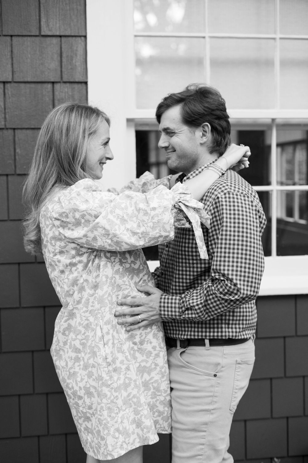 Black and white photo of a smiling pregnant couple embracing outside a shingle-style home.