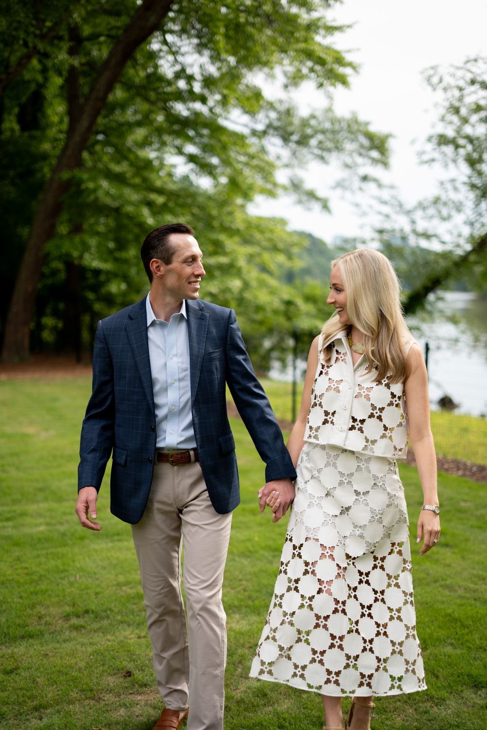Smiling couple holding hands walking in a lush green park near a lake, man in navy blazer, woman in white lace dress.