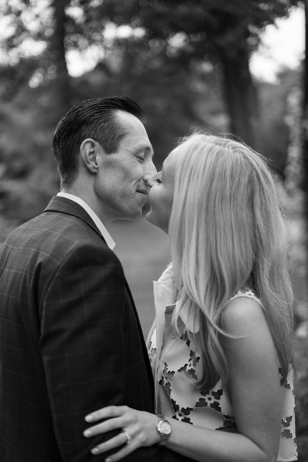 Black and white photo of a couple sharing a tender kiss outdoors among trees.