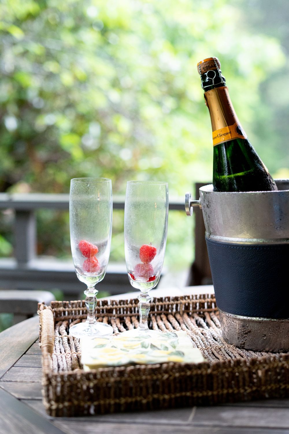 Champagne bottle in ice bucket with two flutes holding raspberries on a wicker tray.