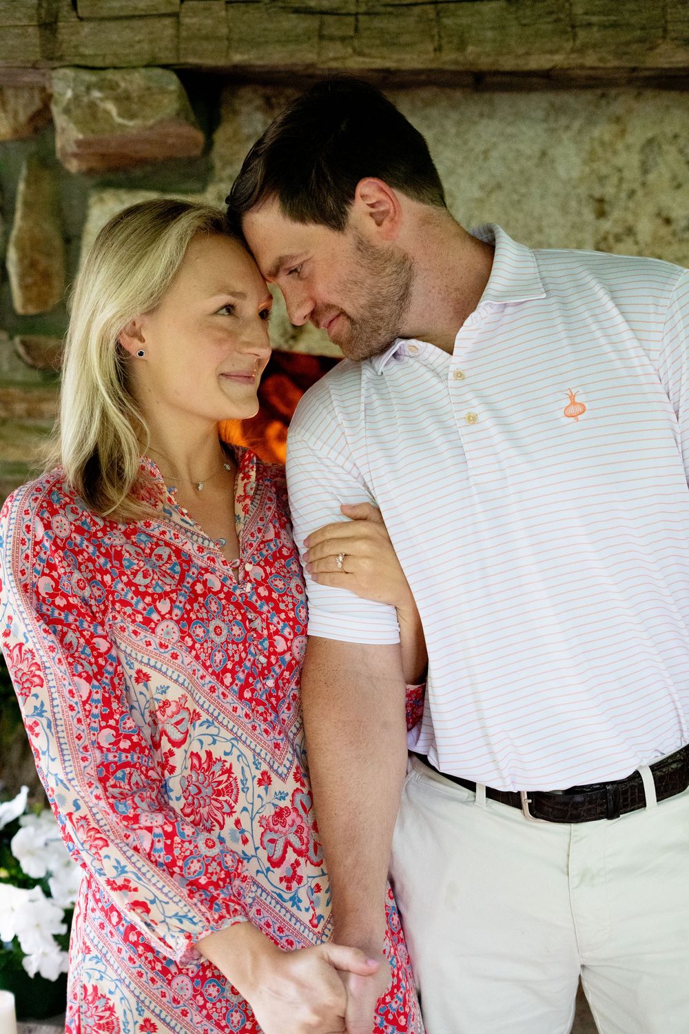 Romantic couple embracing near a stone fireplace, woman in red floral dress, man in white polo shirt.