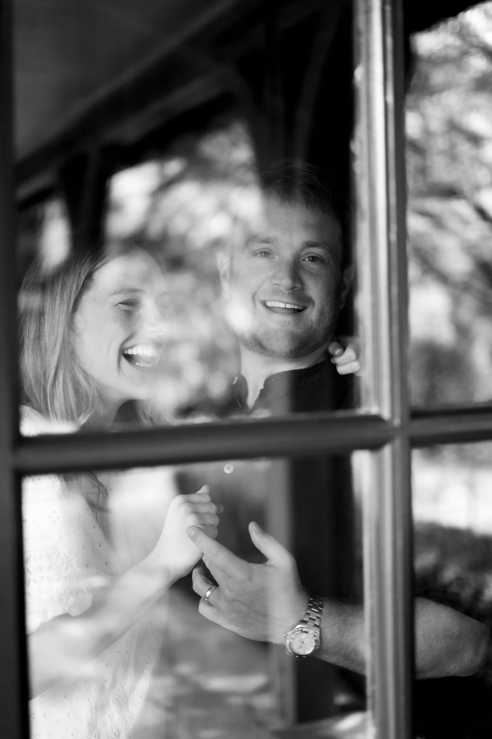 Black and white photo of a joyful couple holding hands and smiling through a window.