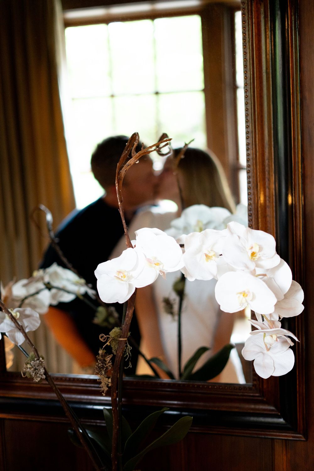 Couple kissing reflected in a mirror behind a blooming white orchid plant indoors.