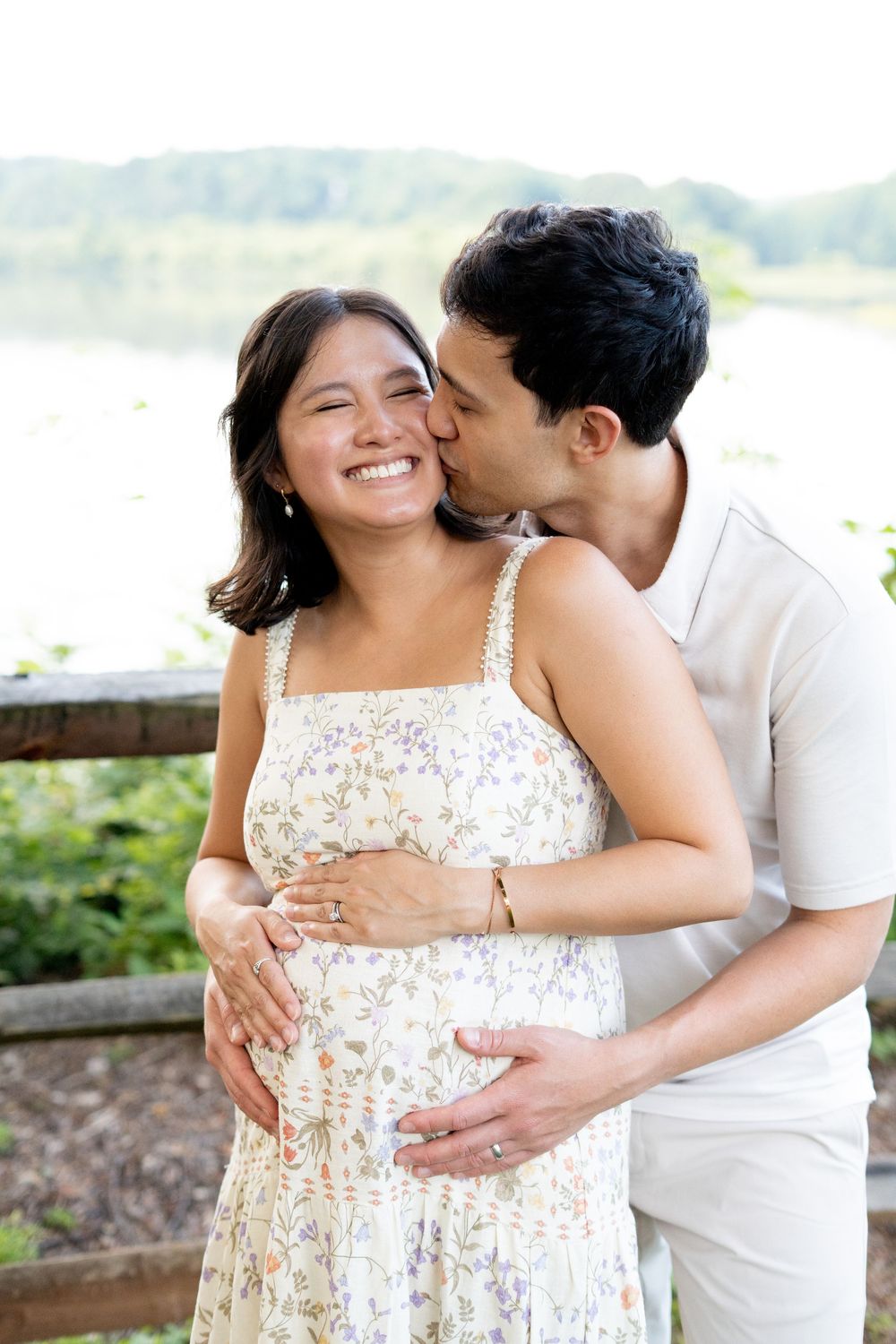 Happy pregnant woman smiling as her partner kisses her cheek near a scenic lakeside backdrop.