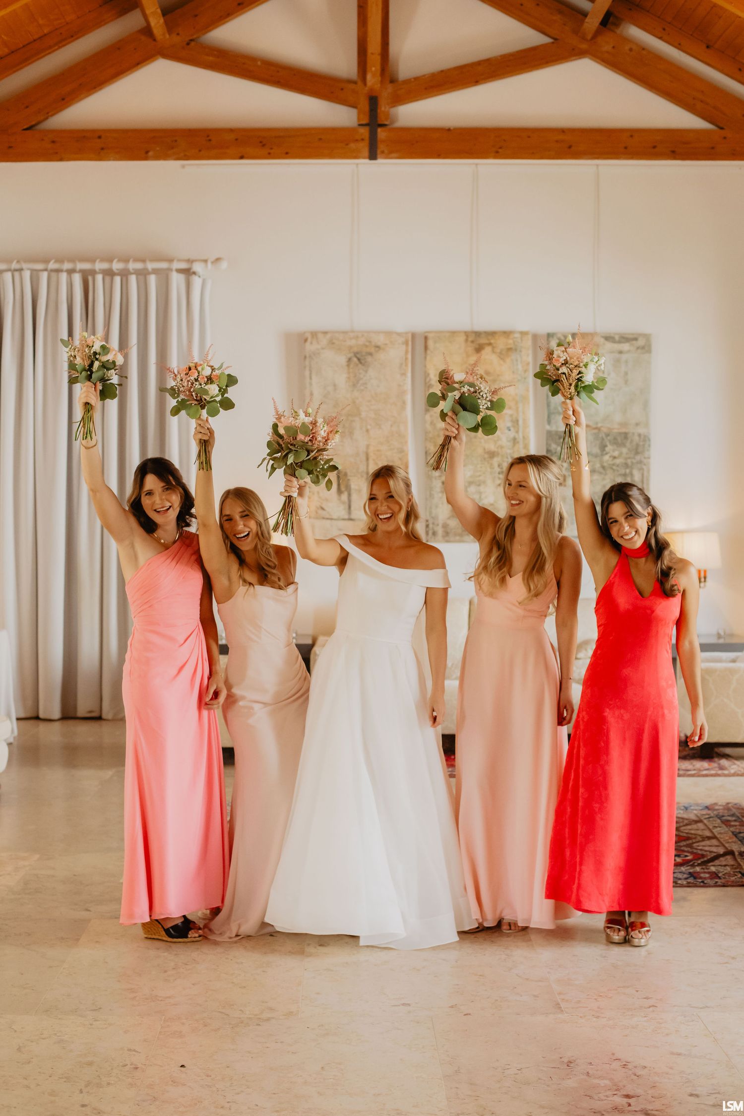 Bride in white gown with four bridesmaids in pink and red dresses raising bouquets indoors.
