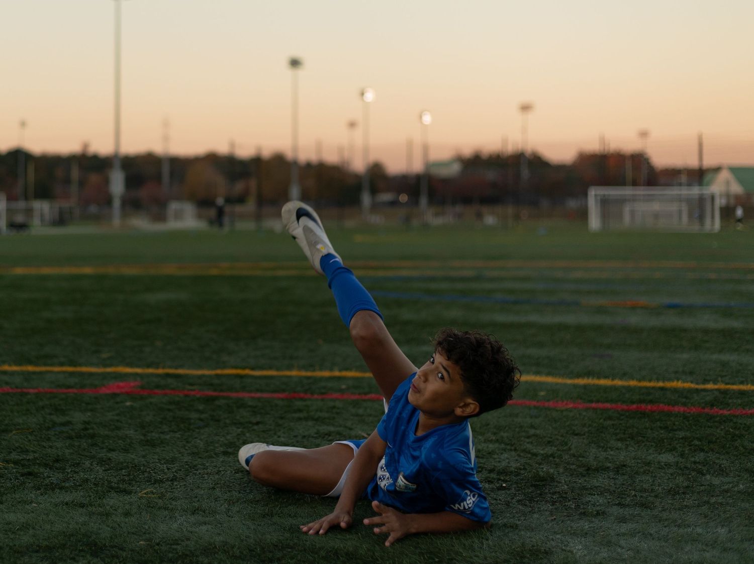 Young soccer player in blue uniform stretching leg up on green field at dusk with goal post in background.