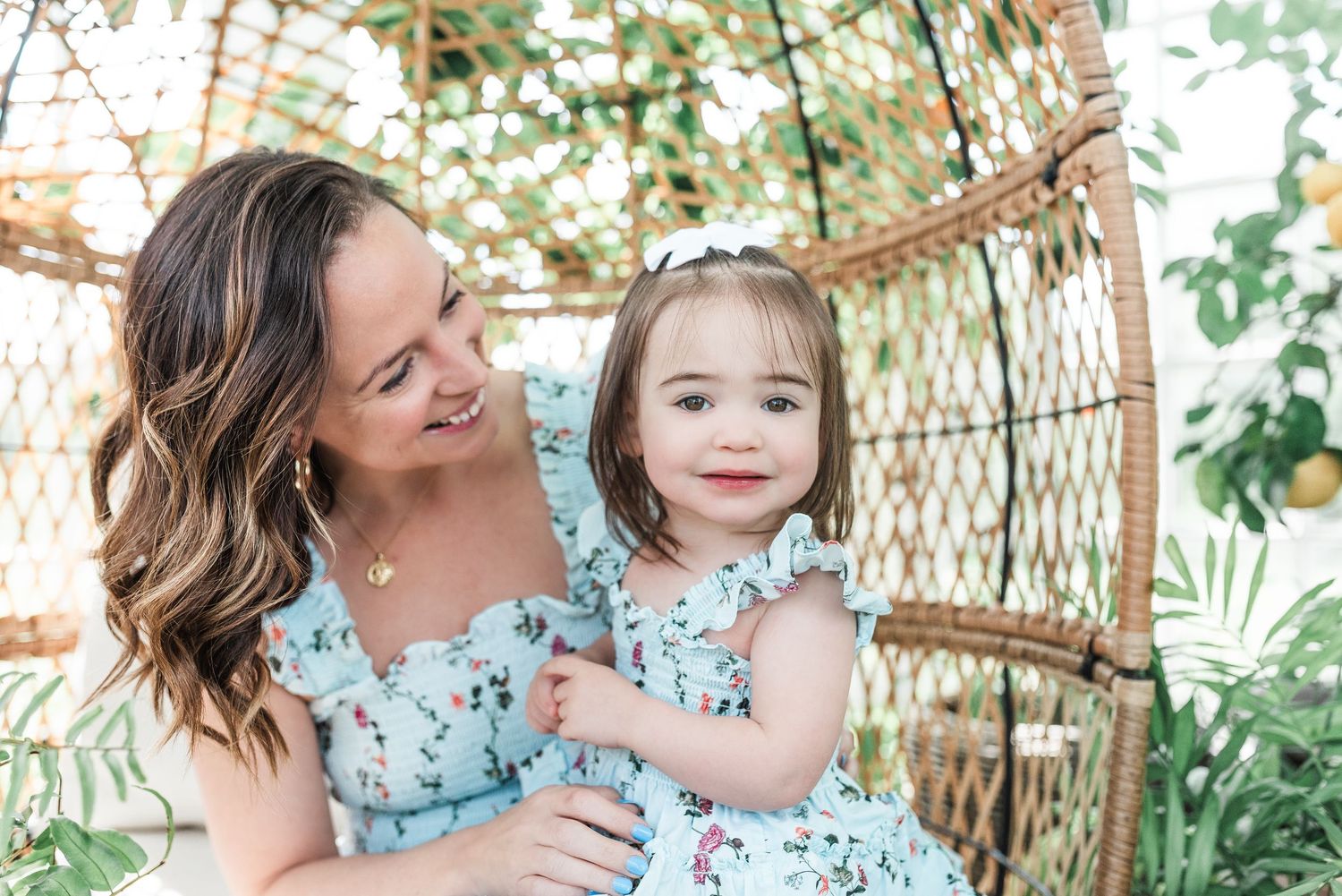 Mother and daughter wearing matching floral dresses sit together in a wicker peacock chair in a garden setting.