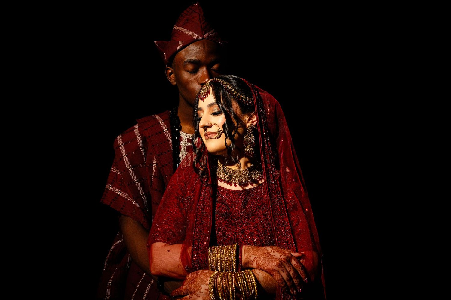 Interracial couple in traditional Indian wedding attire, woman in red bridal lehenga with gold jewelry poses elegantly.