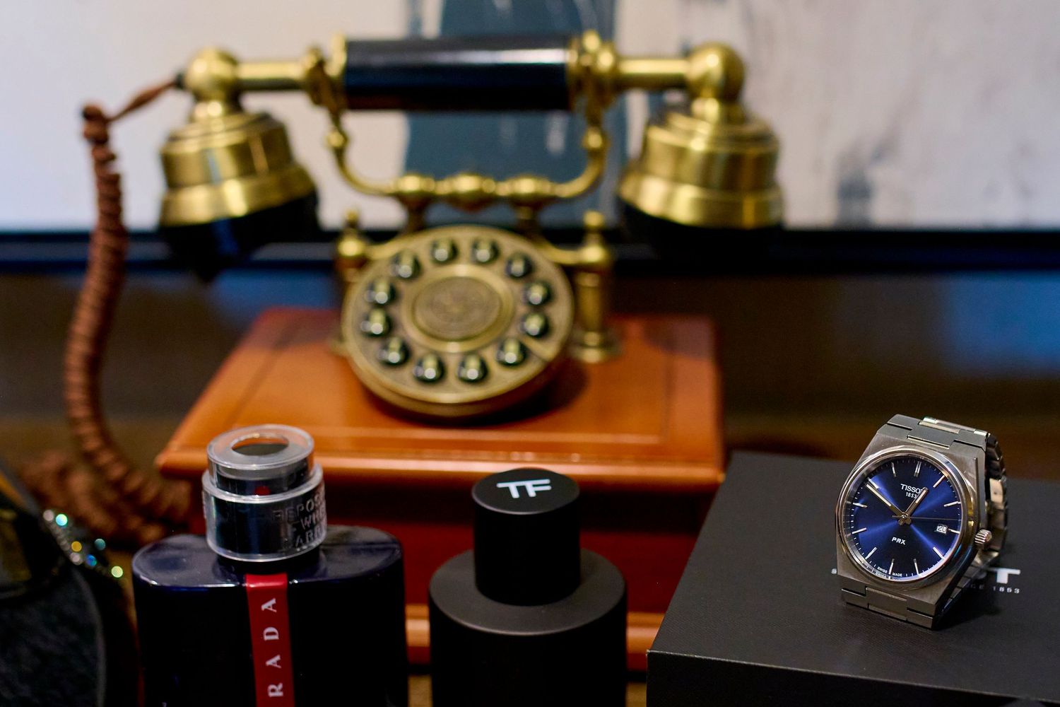 Vintage brass rotary telephone with Prada cologne, Tom Ford perfume, and a blue-dial luxury watch on a dark display surface.