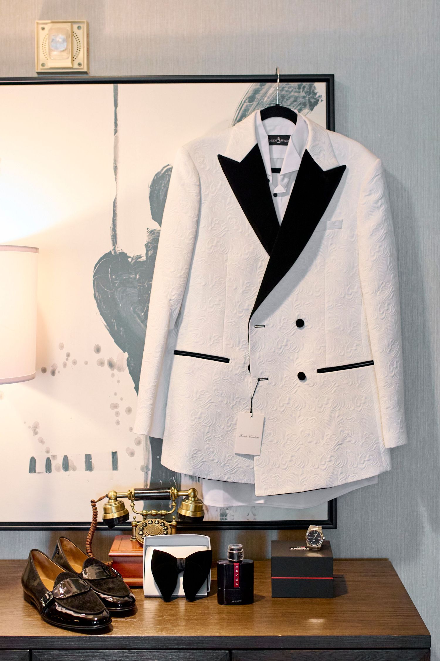 White tuxedo jacket with black lapels on hanger, displayed above loafers, sunglasses, and accessories on wooden dresser.