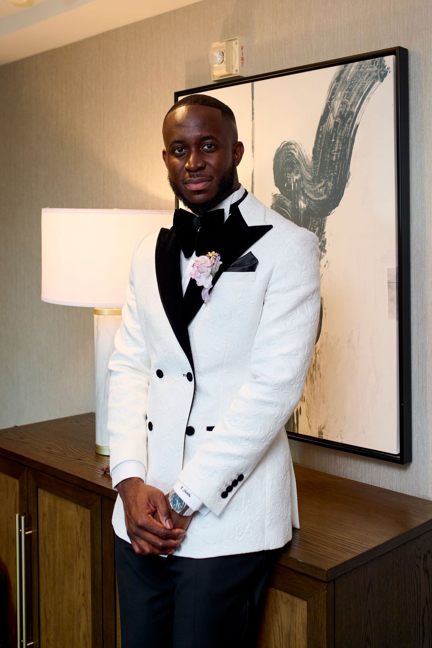 Elegant man in white tuxedo jacket with black lapels and pink boutonniere posing indoors.