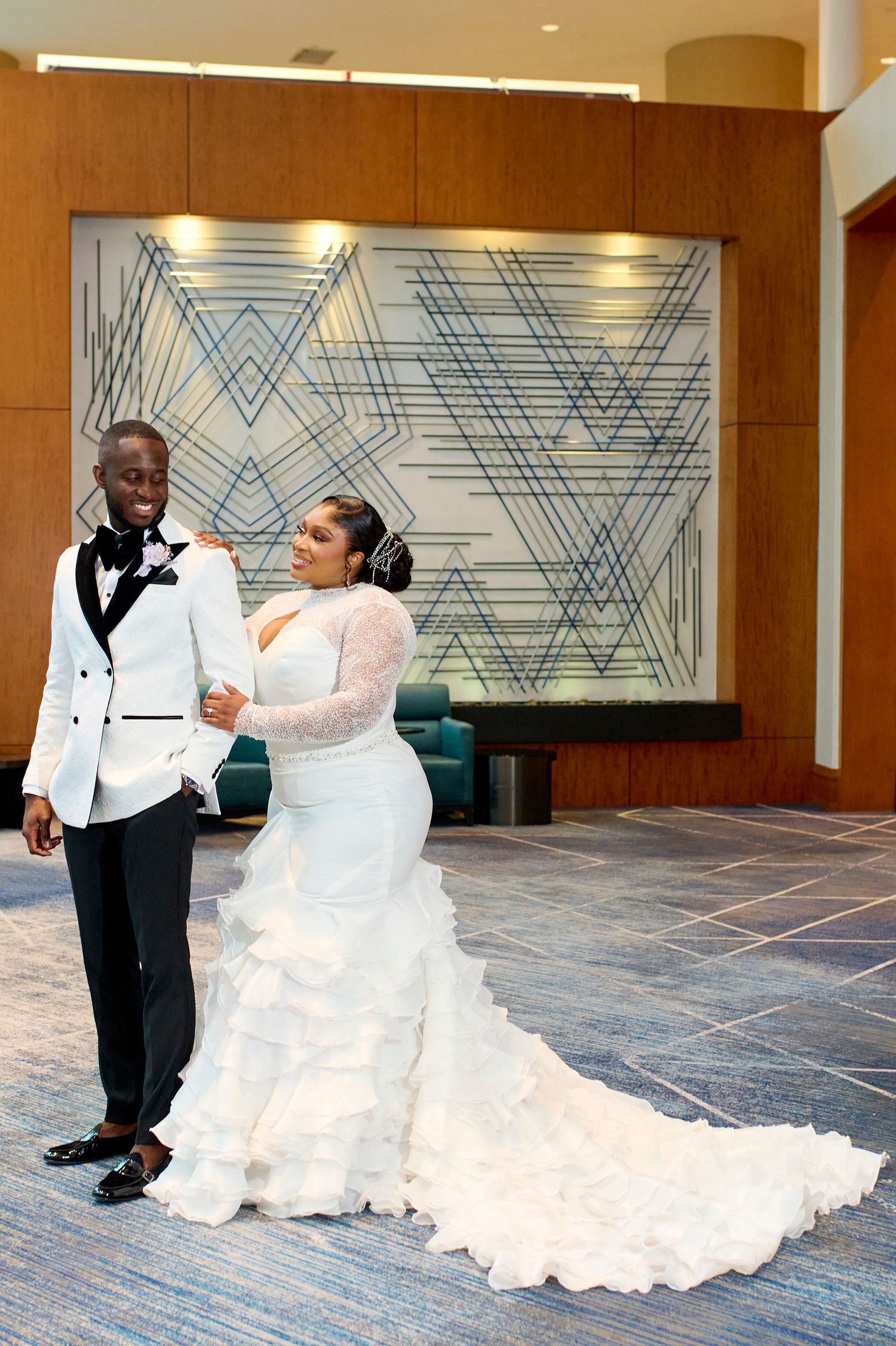 Black couple in wedding attire, groom in white tuxedo jacket, bride in ruffled mermaid gown, posing indoors.