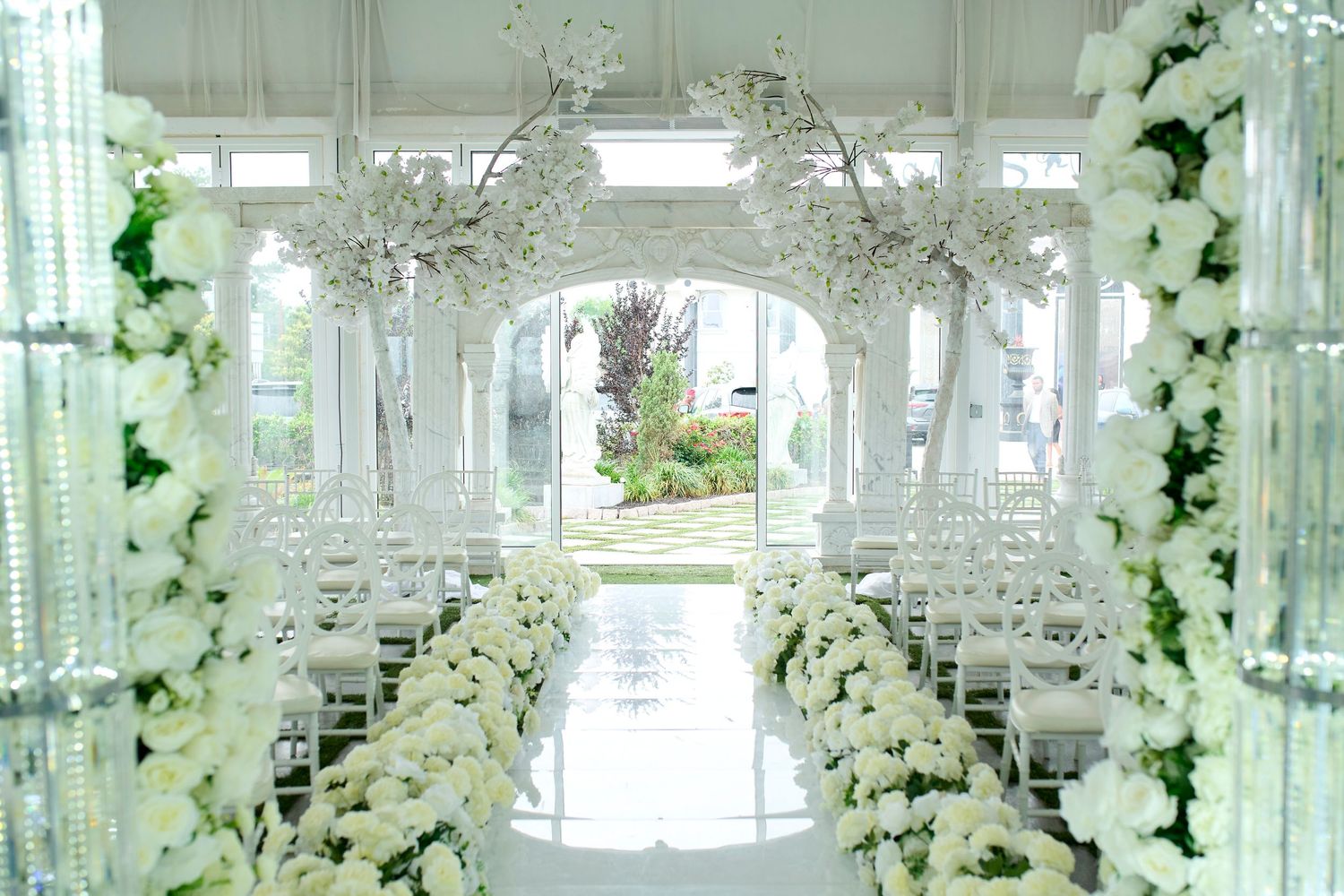 Elegant white wedding ceremony aisle with floral arch, crystal chandeliers, and white chairs in a bright venue.