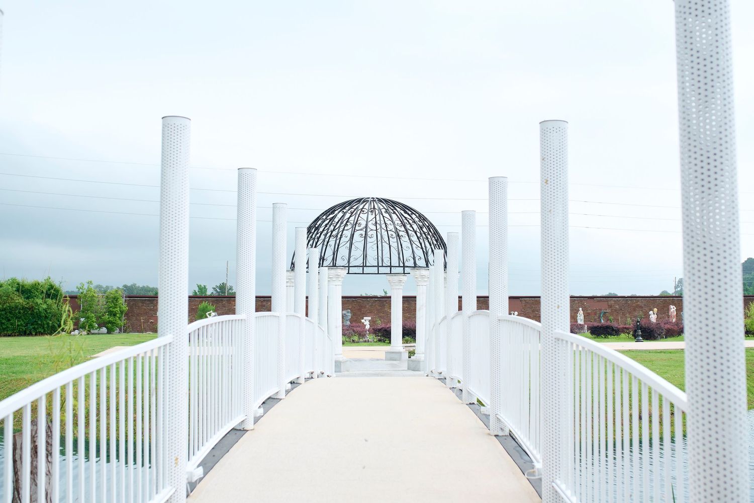 White bridge leading to elegant domed gazebo with tall columns in a lush green park under overcast skies.