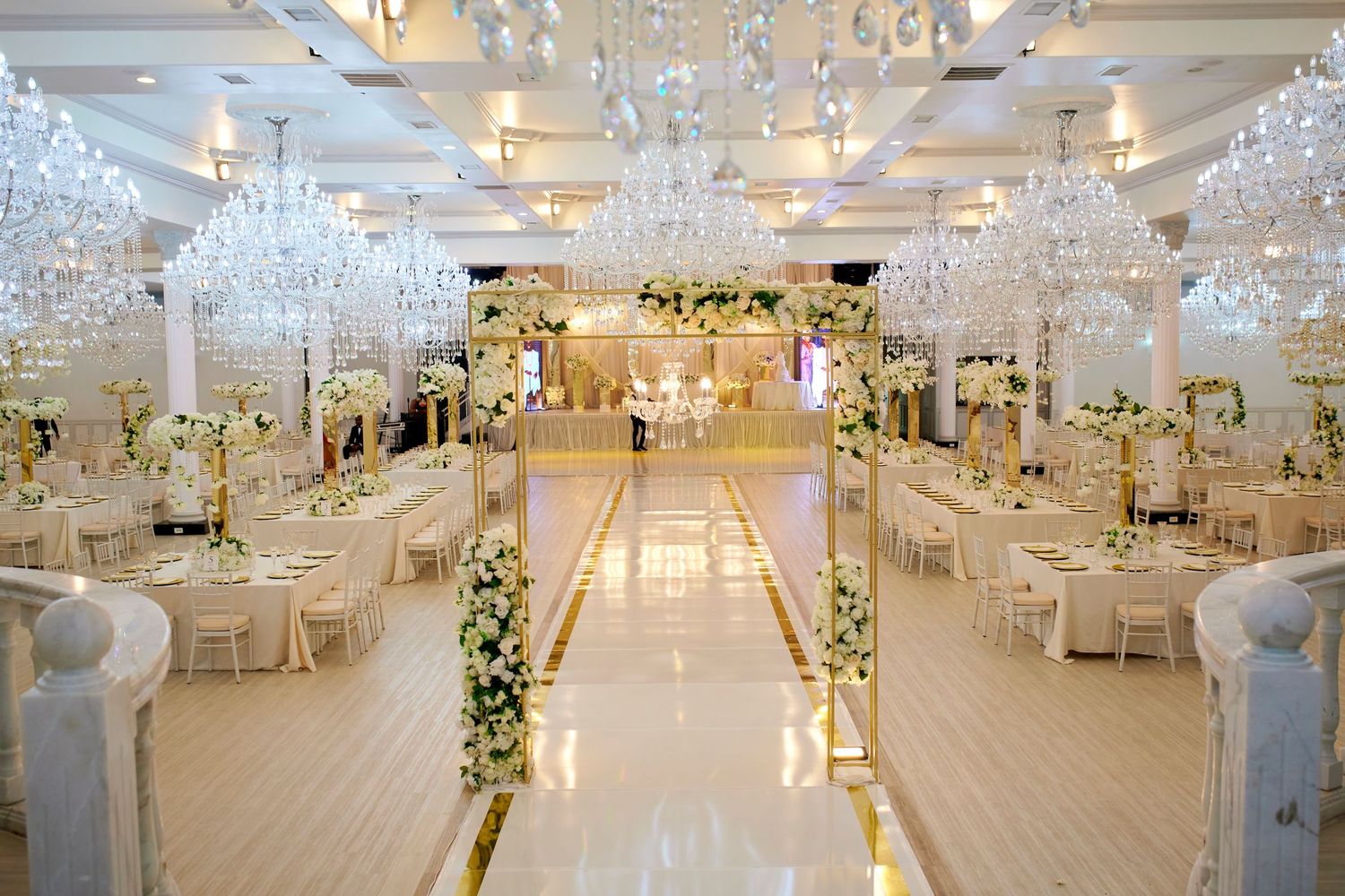 Elegant wedding reception hall with crystal chandeliers, white floral arrangements, gold accents, and a grand aisle leading to the altar.