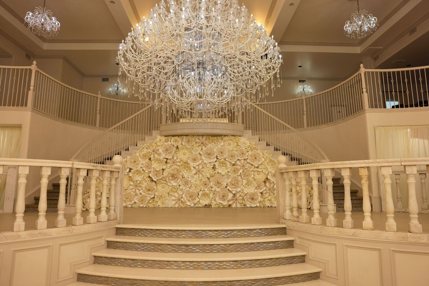 Elegant grand staircase with white floral wall, ornate gold railings, and a large crystal chandelier in a luxurious ballroom.