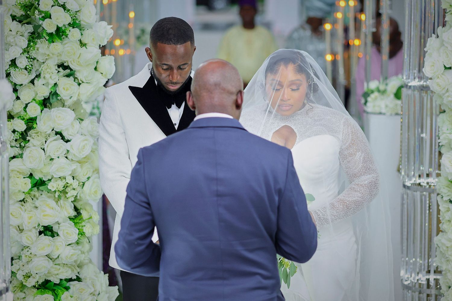 Elegant wedding ceremony with bride in white gown and groom in white tuxedo bowing before officiant amid floral decor.