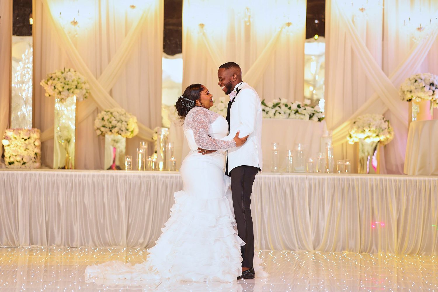 Bride and groom share first dance in elegant white wedding reception venue with floral arrangements and draped backdrop.