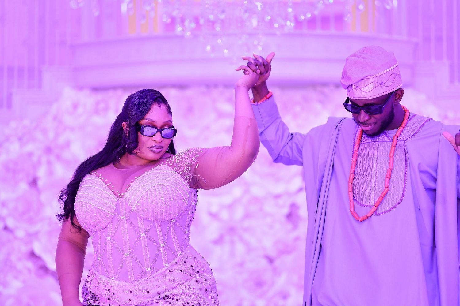 Couple holding hands at elegant purple-lit wedding reception, woman in beaded corset dress, man in traditional Nigerian attire.