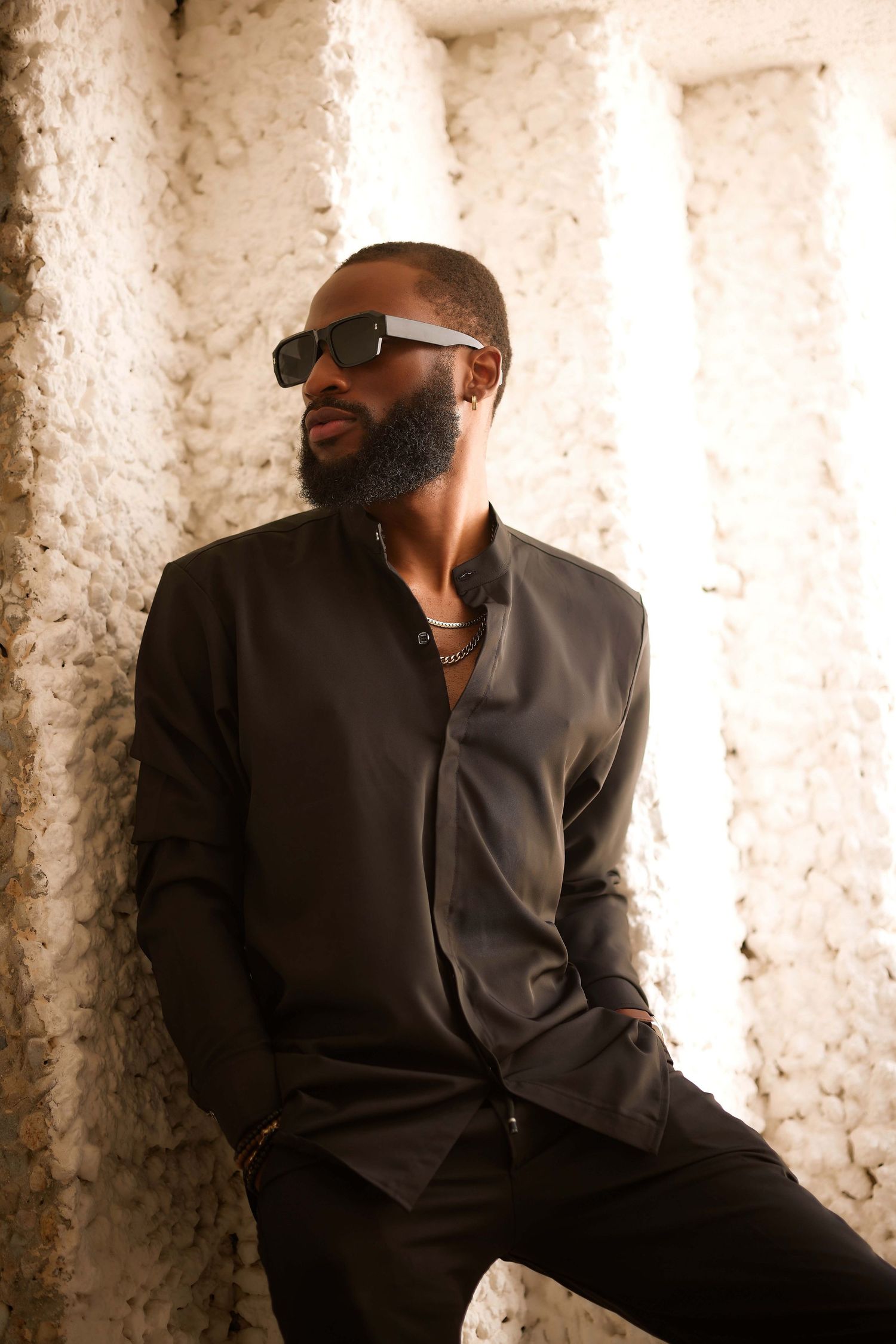 Stylish Black man in dark shirt and sunglasses poses confidently against textured white wall in fashion portrait.