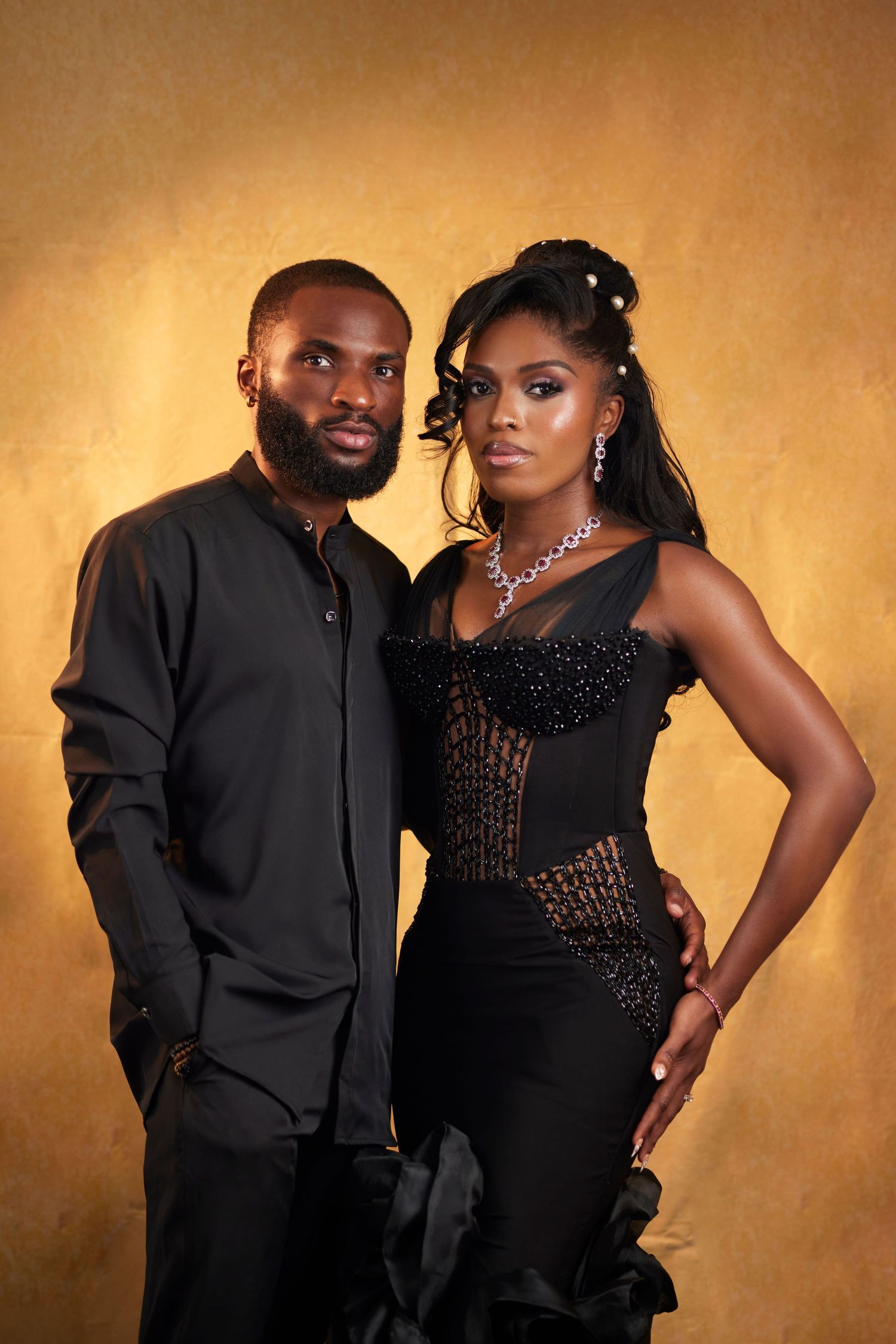 Elegant Black couple posing in formal black attire against golden background, woman wearing diamond jewelry.