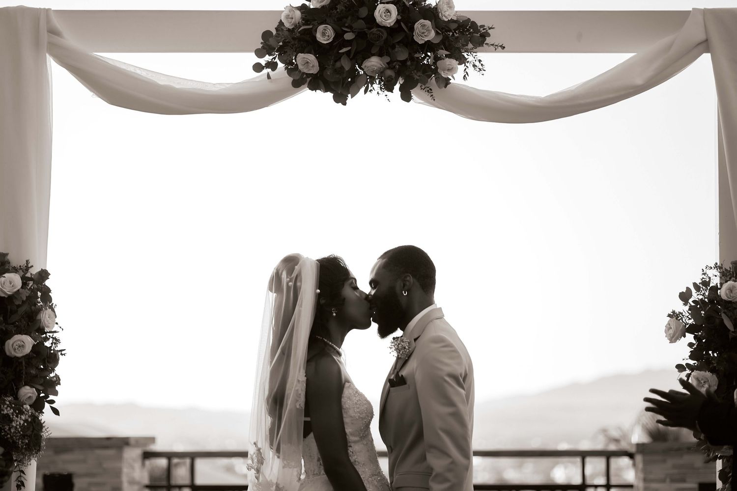 Black and white photo of a bride and groom sharing a kiss under a floral arch at their wedding ceremony.