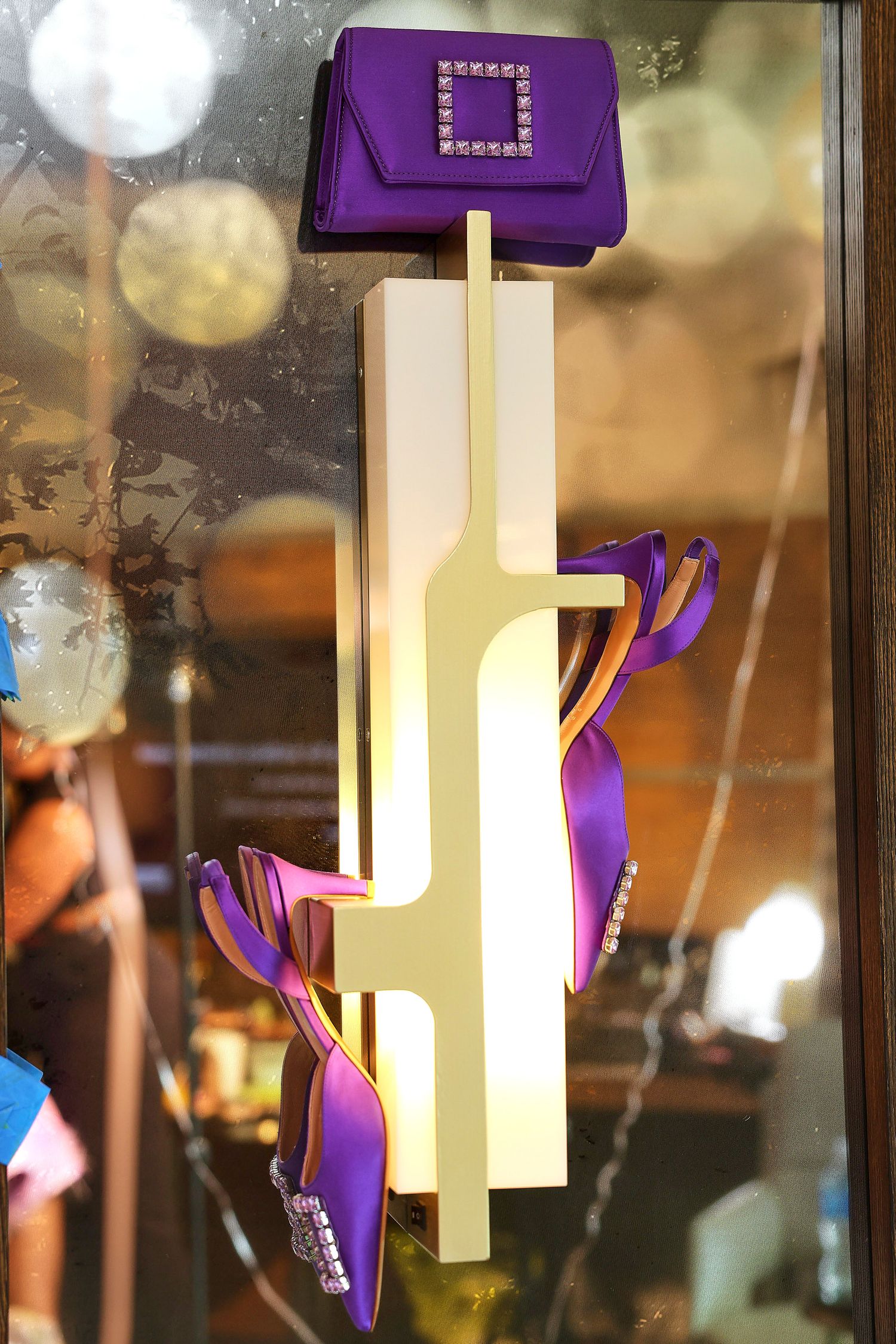 Purple crystal-embellished clutch bag and matching heels displayed on a gold illuminated stand in a luxury store window.